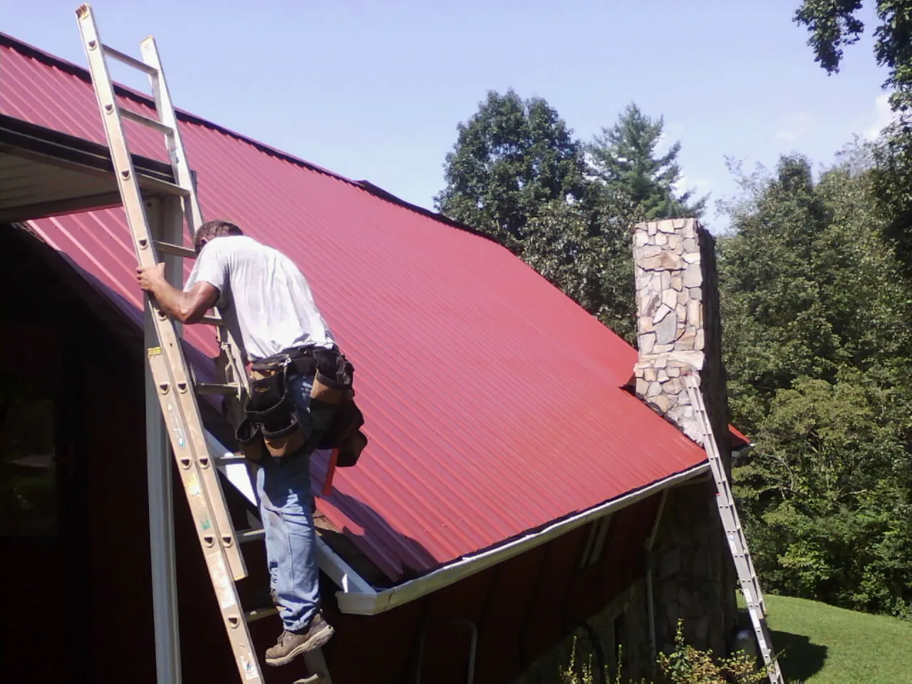 Roofer on a ladder beside a red metal roof and stone chimney — Soffit & Fascia Repair by Roofing Brothers in Harbor Springs